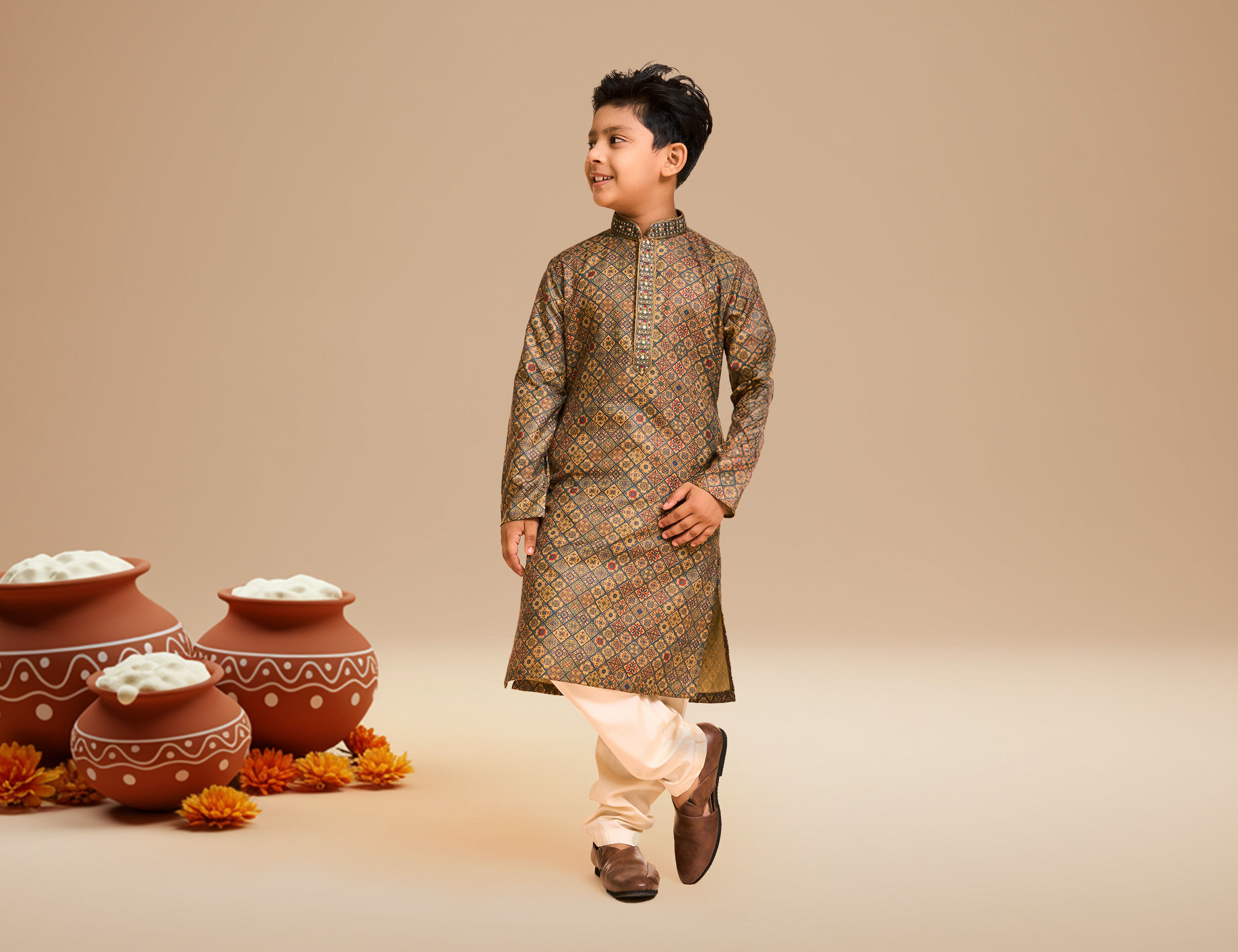 Smiling young boy in traditional brown printed kurta pajama for Janmashtami celebration, standing beside decorated pots filled with butter.